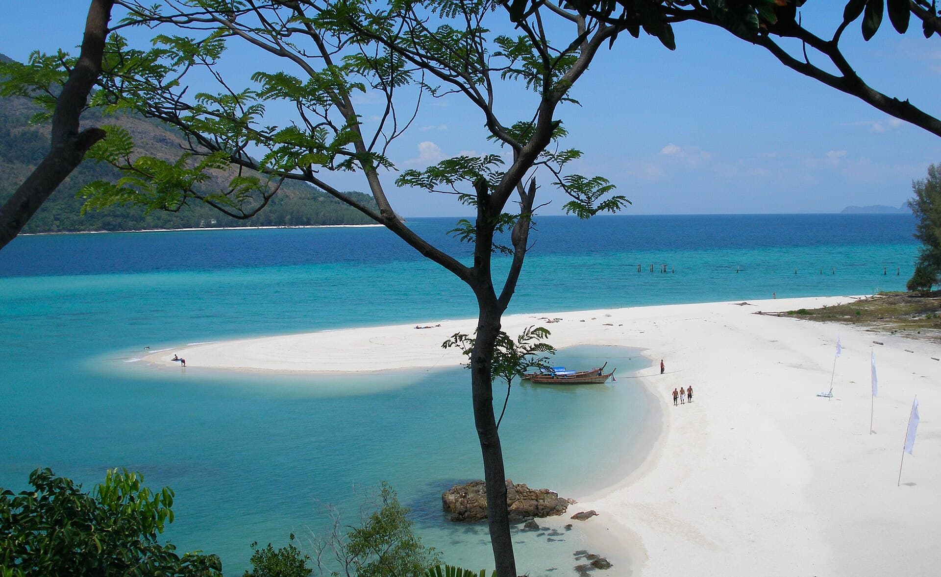 A white sandbar, a longtail boat and turquoise water at Koh Lipe, with Koh Adang behind