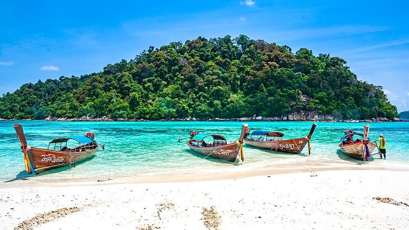 A row of longtail boats moored on a white beach below a forested island